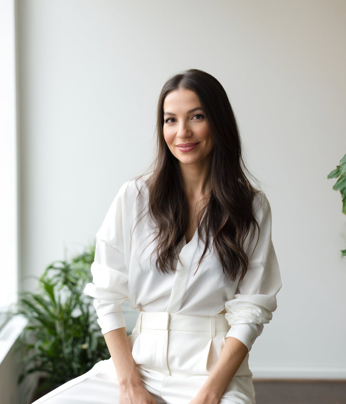 Aline Delamare sorrindo, de camisa e calça brancas, em um ambiente claro com plantas